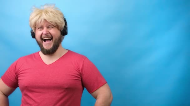 Γενειοφόρος άνδρας με αστείο χτένισμα σε ροζ T-shirt και ακουστικό χαίρεται και γελάει στο μπλε backgound — Αρχείο Βίντεο