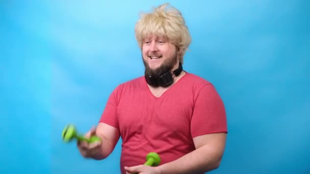Hombre divertido monstruos en los auriculares y con un corte de pelo de 70 pecado rosa camiseta trata de pequeñas mancuernas verdes y sonrisas — Vídeos de Stock