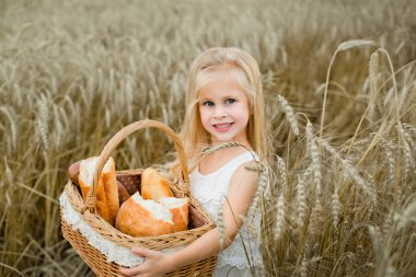 Bir kız elinde bir sepet, ekmek, çörek, un ürünleri, bir simit, mısır gevreği ekmeğiyle, yaz aylarında, gün batımında, beyaz bir elbise içinde bir buğday tarlasında duruyor.