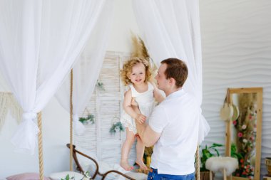 Baba neşeyle gülen küçük kızını kusar. Evde, yatak odasında oynarken mutlu ol. Babası küçük kızını kollarında tutuyor..