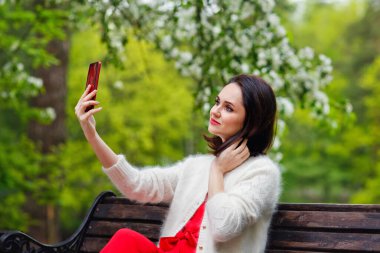 Bir model parkta gülümseyerek poz veriyor, açık hava bankında oturuyor ve elinde akıllı bir telefonla konuşuyor. Harika makyajlı güzel esmer bir kızın portresi. Bir kız telefonuyla kendi fotoğrafını çekiyor.