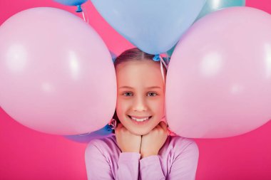 Renkli balonları olan güzel bir kız pembe arka plana gülüyor. Yüzüne balon yapıştırmış bir kızın büyük bir portresi. Pastel renkli bir balonla oynar ve kutlar.