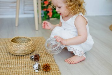 Küçük bir kız gülüyor ve çam kozalağıyla oynuyor. Şirin, genç, sarışın bir kız sınıfta Montessori tarzı ya da ev konsepti ile ders çalışıyor. Doğal maddeleri keşfeder..