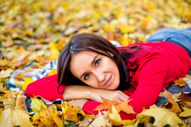 Koyu renk saçlı, kırmızı kazaklı bir kadın sarı akçaağaç yapraklarının arasında bir parkta bir battaniyenin üzerinde yatıyor. Doğada sonbahar pikniği.