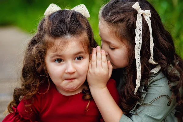 Two little girls with long hair in a red and green dress are sitting, playing and having fun in the Park on the path. Two happy sisters in nature. Says a secret in your ear.