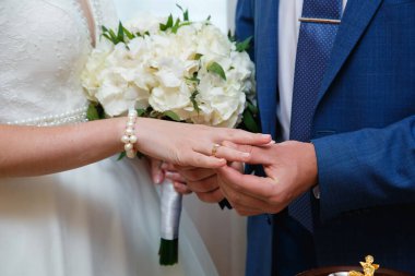 Damat, gelinin parmağına alyans takıyor. Gelin ve damat düğün töreninde el ele tutuşuyorlar. Evlilik yüzükleri yeni evlilerin ellerinde..