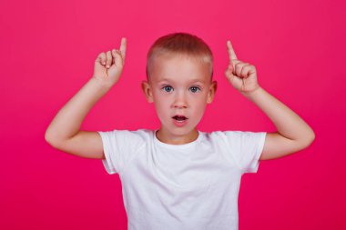 Mavi gözlü, pembe arka planda beyaz tişört giyen ve başarılı bir fikirle parmağını kaldıran küçük beyaz bir beyaz adam. Heyecanlı ve mutlu