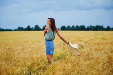 Elinde şapkası olan zayıf bir kız gökyüzünde bulutlarla buğday tarlasının ortasında dönüyor..