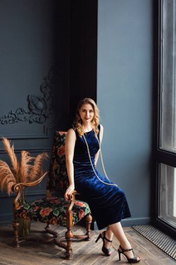 Mavi kadife elbiseli ve cam ve sandalyenin yanında inci boncuklu güzel genç bir kadın..