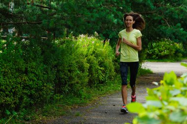 Sarı spor bluzlu ve taytlı güzel bir fitness kızı parkta bir patika boyunca koşuyor. Sıcak bir yaz gününde hızlı koşar. Sporcu kadın her zamanki spor antrenmanına başladı..