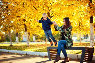 Güzel anne ve oğul sonbahar parkında bir bankta oynuyorlar. Oğlan banktan atlar. Ebeveynler ve çocuklar arasındaki ilişkiler. Anneler Günü.