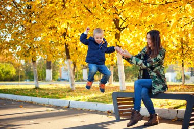 Güzel anne ve oğul sonbahar parkında bir bankta oynuyorlar. Oğlan banktan atlar. Ebeveynler ve çocuklar arasındaki ilişkiler. Anneler Günü.