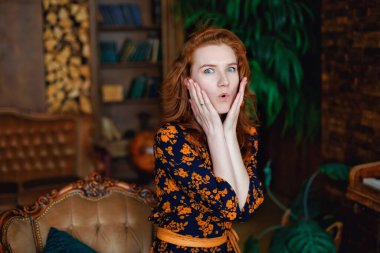 Güzel, kızıl saçlı genç bir kadının portresi, çok şaşırmış, kameraya bakıyor ve ellerini yüzüne dayıyor.