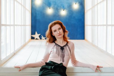 Çekici kızıl saçlı genç bir kadın beyaz pencerenin yanındaki geniş bir odada poz veriyor ve mavi bir duvarda çelenk var..