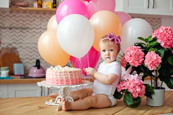 A little girl laughing sits on a table with a birthday cake and balloons. Celebration of the first year.