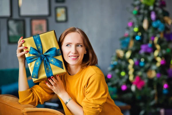 Sarı kazaklı, kızıl saçlı bir kadın Noel ağacının arka planında kulağına altın sarısı bir hediye tutuyor..