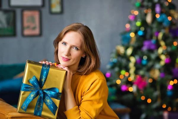 Sarı kazaklı kızıl saçlı bir kadın bir Noel ağacının arka planına sarılan bir hediye tutuyor.
