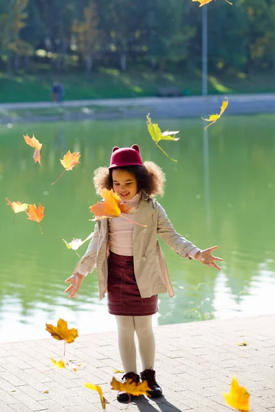 一个女孩在湖边玩落叶游戏 迷人的女学生放学后在秋天的公园散步 一个黑皮肤的孩子扔秋叶 生活方式 — 图库照片