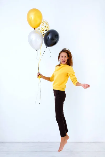 Una mujer joven en un suéter amarillo salta y sostiene un montón de globos en oro, blanco y plata contra una pared blanca. — Foto de Stock