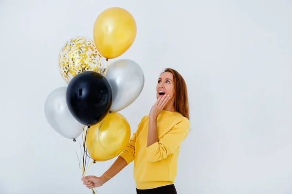 Een jonge vrouw in een gele trui houdt een bos gouden, witte en zilveren ballonnen tegen een witte muur. Hij kijkt verbaasd naar de ballen. Ruimte voor tekst — Stockfoto