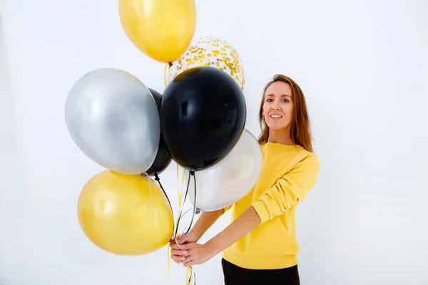 Een jonge vrouw in een gele trui houdt een bos gouden, witte en zilveren ballonnen tegen een witte muur. — Stockfoto