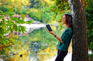 Genç, siyah saçlı bir kadın, elinde bir kitapla gölün yakınındaki bir ağaca yaslanıyor..