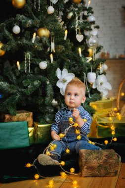 Küçük gülümseyen bir çocuk, bir Noel ağacının arka planında bir bebek, elinde ışıklardan bir çelenk ve kurdeleli bir hediye kutusuyla.