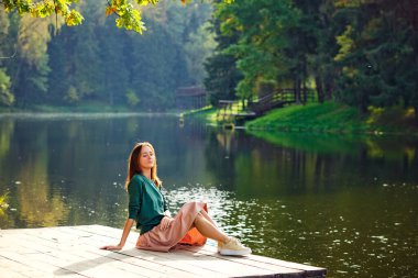 Arka planda nehir ve ormanın rıhtımında oturan rüya gören genç bir kadın..