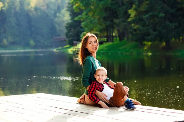 A beautiful mother sits with her son on the edge of the lake bridge, her legs dangling down, with a beautiful landscape of nature.