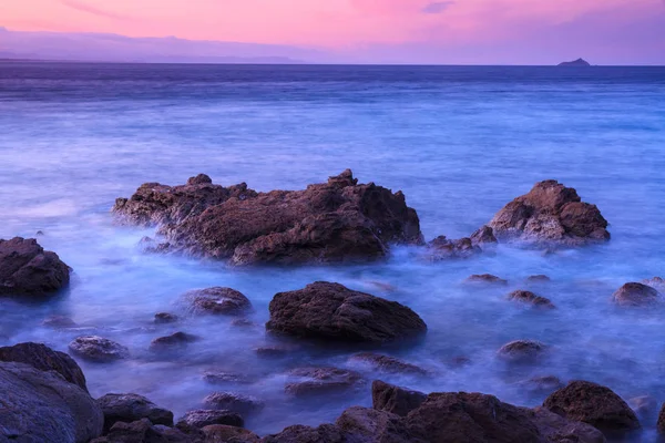Rocky shore under purple skies at dusk. A long exposure has turned the water breaking over the rocks into a misty haze