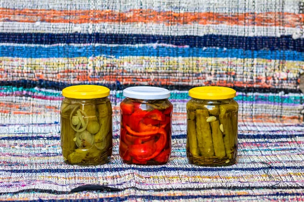 Glass jars with pickled red bell peppers and pickled cucumbers (pickles) isolated. Jars with variety of pickled vegetables. Preserved food concept in a rustic composition.