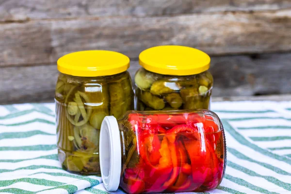 Glass jars with pickled red bell peppers and pickled cucumbers (pickles) isolated. Jars with variety of pickled vegetables. Preserved food concept in a rustic composition.