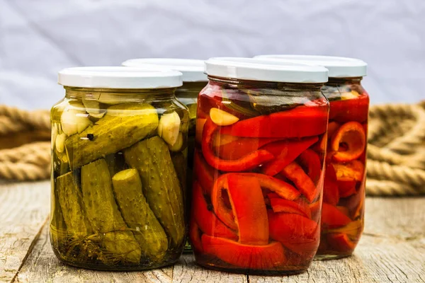 Glass jars with pickled red bell peppers and pickled cucumbers (pickles) isolated. Jars with variety of pickled vegetables. Preserved food concept in a rustic composition.