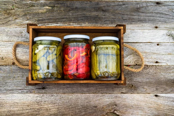 Glass jars with pickled red bell peppers and pickled cucumbers (pickles) isolated in wooden crate. Jars with variety of pickled vegetables. Preserved food concept in a rustic composition.