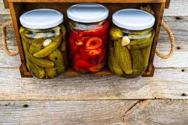 Glass jars with pickled red bell peppers and pickled cucumbers (pickles) isolated in wooden crate. Jars with variety of pickled vegetables. Preserved food concept in a rustic composition.