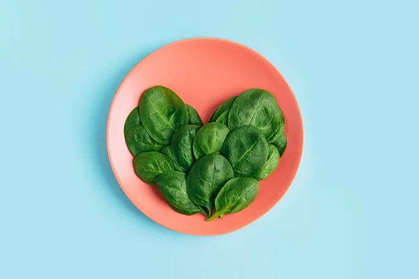 Green fresh vegetarian salad leaves shaped heart on coral plate on blue background.