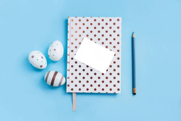 Notebook pontilhado branco, pecil, ovos decorativos de Páscoa e papel branco sobre fundo azul . — Fotografia de Stock