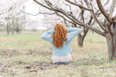 Beyaz elbiseli ve mavi kazak lı romantik genç kadın, ilkbahar çiçek açan bahçede yerde oturuyor. Geri görünüm.