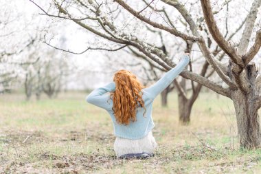 Beyaz elbiseli ve mavi kazak lı romantik genç kadın, ilkbahar çiçek açan bahçede yerde oturuyor. Geri görünüm.