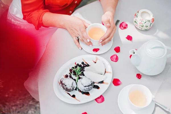 Teetassen und Frauenhände, die eine Tasse auf einem Tisch mit Rosenblättern halten, Ansicht von oben. — Stockfoto