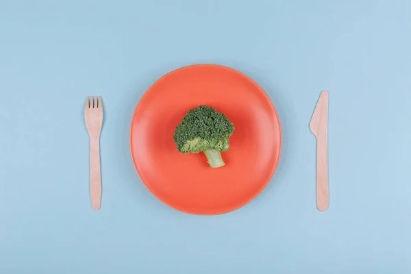 Маленький кусочек свежей брокколи на оранжевой тарелке с деревянной вилкой и ножом . — стоковое фото
