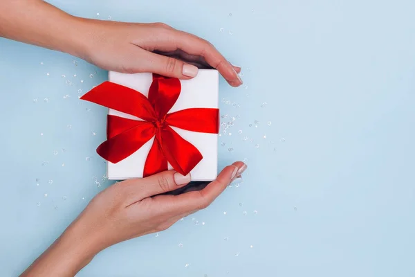 Frauenhände mit Maniküre halten Geschenkbox mit roter Schleife auf hellblauem Hintergrund mit Glitzern. Festliches Konzept. — Stockfoto