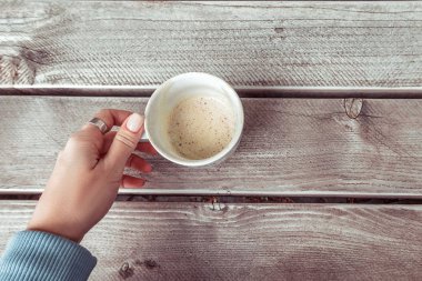 Mavi kazaklı bir kadın, gri ahşap masada boş bir fincan kahve tutuyor..