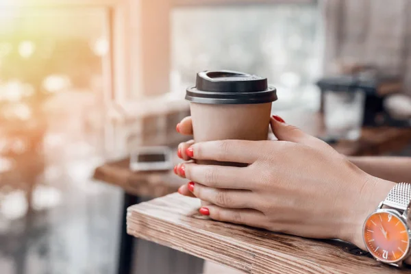 Paper coffee cup to go in womans hands with red manicure while sitting in cafe.
