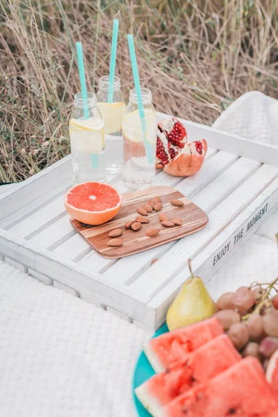 Picknickset mit Grapefruit, Granatapfel, Mandelnüssen, Wassermelone, Trauben und Limonade in Glasflaschen auf weißem Tablett. — Stockfoto