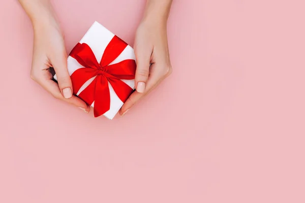 Las manos de la mujer con manicura sostienen la caja de regalo con cinta roja sobre un fondo rosa pálido. Concepto festivo . — Foto de Stock