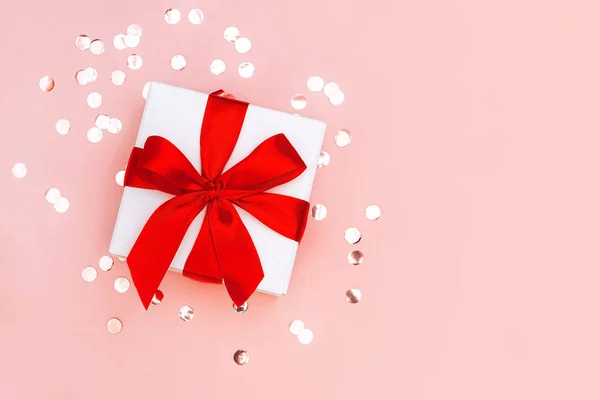 Caja de regalo con cinta roja sobre fondo rosa pálido con confeti dorado. Concepto festivo . — Foto de Stock