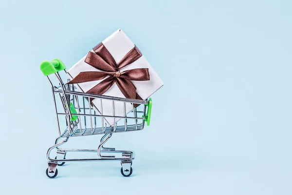 Gift box with brown bow in miniature shopping cart on pastel blue background with copy space.