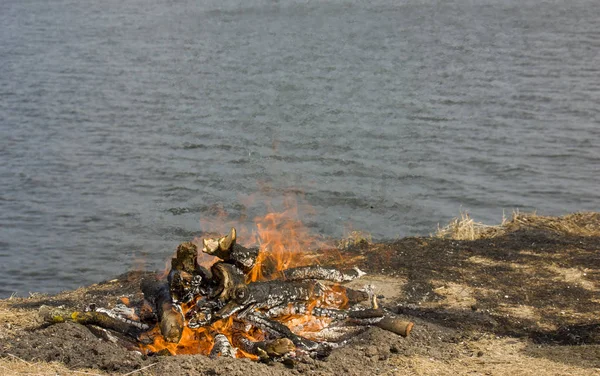 A large bonfire with fire sparks of flame on the banks of the river. Burned grass by the fire. Close-up.