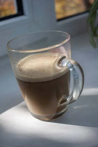 Delicious aromatic morning coffee with milk in a glass mug with double walls on a windowsill in the sunshine